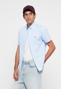 Lys blå kortermet skjorte med knapper, hvit t-skjorte under, lys blå denimjeans. Modellen har på seg en burgunder caps med logo.