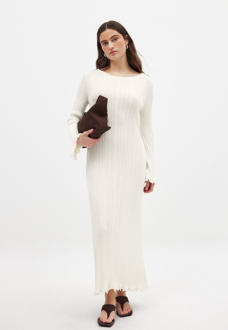 Lång, ribbad klänning i vitt med utsvängda, scallopade ärmar och scallopad nederkant. Modell som håller en brun, texturerad clutch. Flip-flops i mörkbrunt.