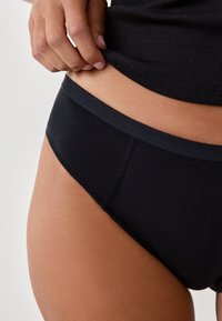 Zwarte bikini-bodem met een gladde textuur, lage taille en een naadloze tailleband, iets omlaag geschoven bij de heup.