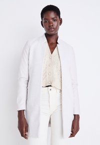 Cappotto grigio chiaro con un colletto strutturato, realizzato in un tessuto liscio. Indossato sopra un top in maglia corto color crema e pantaloni bianchi a vita alta.