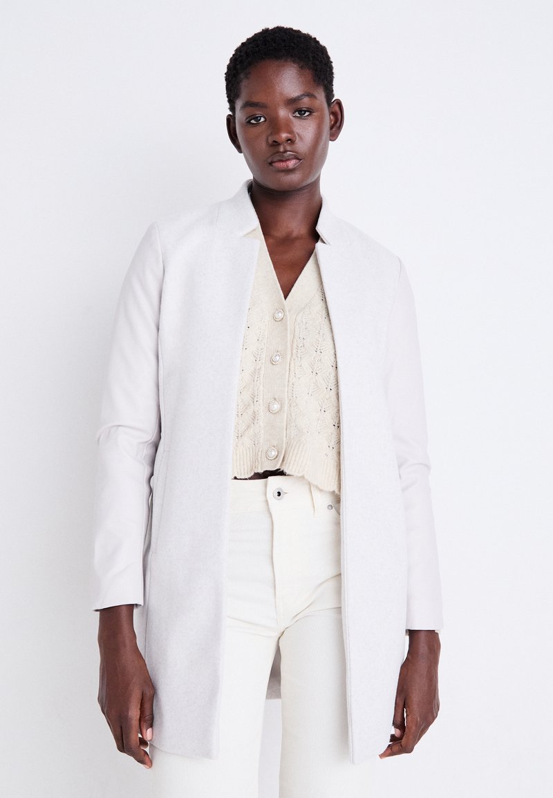 Cappotto grigio chiaro con un colletto strutturato, realizzato in un tessuto liscio. Indossato sopra un top in maglia corto color crema e pantaloni bianchi a vita alta.