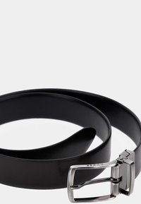 Cintura in pelle nera con una texture liscia e una fibbia in metallo argentato caratterizzata da un design semplice. La cintura è arrotolata e poggia piatta.