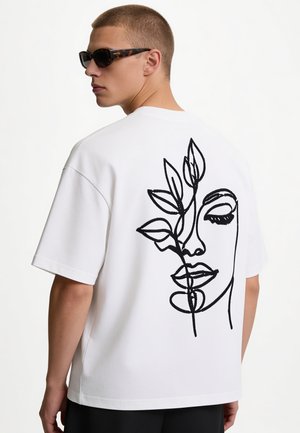 Jonge man met schildpaddensolbril en een wit T-shirt met een zwart lijntekening van een gezicht en bladeren op de achterkant.