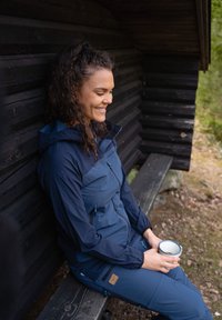 Navy blue outdoor jacket and pants with zippers and blue accents. Sitting on a wooden bench, holding a white cup. Natural backdrop.