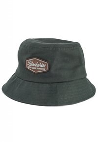 Zelen klobuk iz bombaža v obliki vedra z našitim rjavim oznako, ki vsebuje besedilo "Blackskies" in "HIGH GRADE SUPPLIES". Mehka tekstura, širok rob.