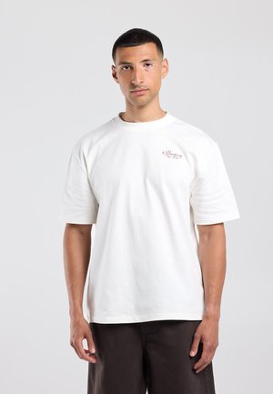 Junger Mann mit kurzen Haaren, der ein weißes oversized T-Shirt und dunkle Hose trägt, steht vor einem schlichten hellen Hintergrund.