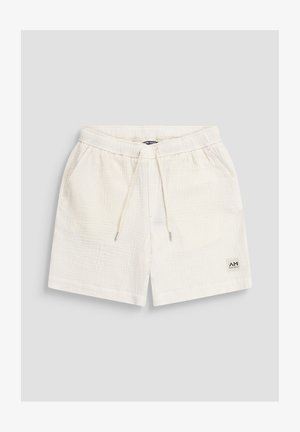 Pantalones cortos de tela blanca con textura, con cintura elástica, cordón ajustable, bolsillos laterales y una pequeña etiqueta de marca en la parte inferior de la pierna izquierda.