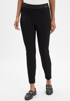 Vrouw gekleed in slim-fit zwarte broek met voornaad en zwarte loafers met gouden gespen, staande tegen een witte achtergrond.