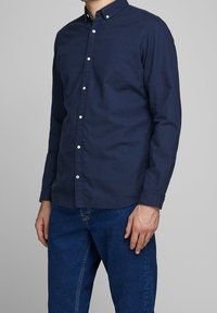 Camisa azul marino abotonada con cuello, mangas largas y botones blancos, combinada con jeans de mezclilla azul. Tela lisa sin patrones visibles.