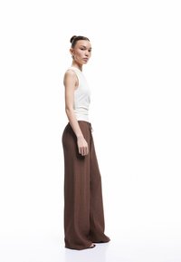 Top blanco sin mangas y pantalones anchos marrones, tejido texturizado, cintura con cordón, modelo de pie de perfil contra un fondo blanco.