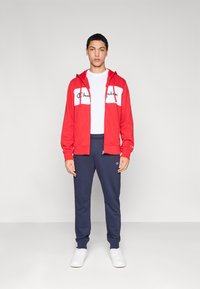 Κόκκινο φούτερ με φερμουάρ και λευκή μπροστινή επιφάνεια με λογότυπο, σε συνδυασμό με σκούρο μπλε αθλητικό παντελόνι και λευκά αθλητικά παπούτσια. Απλός, αθλητικός σχεδιασμός.