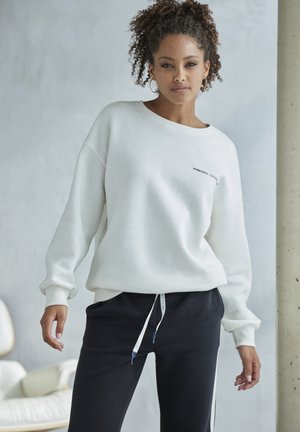 Weißes übergroßes Sweatshirt mit gerippten Bündchen, versehen mit einem kleinen schwarzen Logo. Kombiniert mit enganliegenden schwarzen Sweatpants mit Kordelzug.