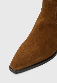 Καφέ σουέτ ankle boot με μυτερή μύτη, διαθέτοντας μαύρη ελαστική σόλα. Η επιφάνεια φαίνεται μαλακή και υφασματώδης, αναδεικνύοντας την ποιότητα κατασκευής της.