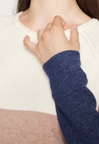 Main avec une manche de pull bleu tirant doucement le col d'un pull en tricot beige et marron clair porté par une personne aux longs cheveux foncés.