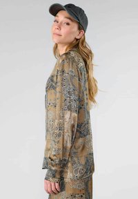 Langsleeve, bruin gebloemd blouse met paisley ontwerpen in zilver en blauw. Zachte stof met een relaxte pasvorm. G gedragen met een donkere pet.