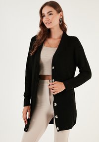 Fekete kötött kardigán hosszú ujjal, V-nyakkal és hat ezüst gombbal. A textúra bordázott, combközépig ér. Bézs felső fölött viselve.