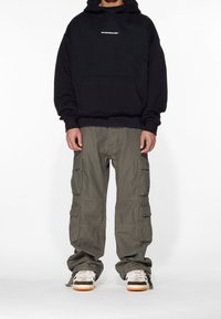 Zwarte hoodie met capuchon en een voorzak, gecombineerd met olijfgroene cargobroek met meerdere zakken, en witte sneakers met bruine accenten.