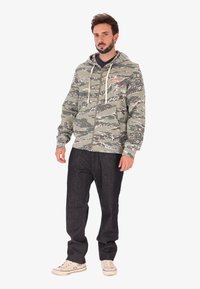 Uomo in piedi che indossa un hoodie verde mimetico con zip, jeans scuri e sneaker bianche su uno sfondo bianco uniforme.