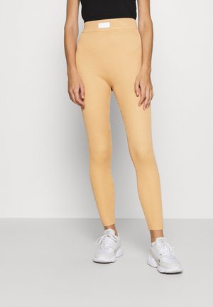 Persona con leggings beige de canalé y zapatillas blancas, de pie contra un fondo claro y liso, con las manos relajadas a los lados.