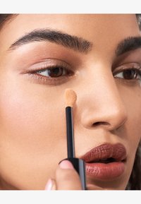 Flytende concealer-applikator i bruk på lys brun hud under øyet. Har en jevn tekstur og en liten, rund svampespiss.