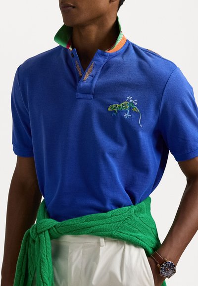 Camisa polo azul com colarinho verde e laranja, apresentando um design de lagarto bordado. Usada com um xaile tricotado verde e calças brancas.