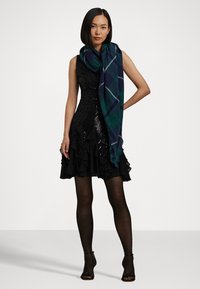 Μαύρο φόρεμα με παγιέτες και φάσα με βολάν, συνδυασμένο με ένα μεγάλο, καρό πράσινο και ναυτικό φουλάρι. Το μοντέλο φοράει διαφανείς μαύρες καλσόν και σχέδια ψηλοτάκουνα.