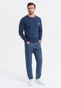 Maglietta a maniche lunghe blu navy con colletto a righe rosse e bianche, indossata con jeans in denim blu e sneakers bianche. Presenta un logo discreto.