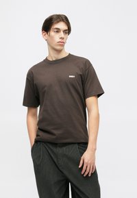 Brun bomulds T-shirt med korte ærmer og rund halsudskæring. Har et lille hvidt "OBEY" logo på venstre bryst.