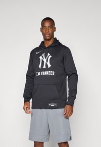 Juodas hoodie su priekinėmis kišenėmis, su baltu „New York Yankees“ logotipu ir „Nike“ ženklu. Dėvimi pilki šortai, pagaminti iš minkštos medžiagos.