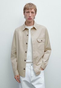 Beige skjorte med knapper lavet af bomuld, med en brystlomme, brune knapper og lange ærmer, parret med en hvid T-shirt med rund hals og hvide bukser.