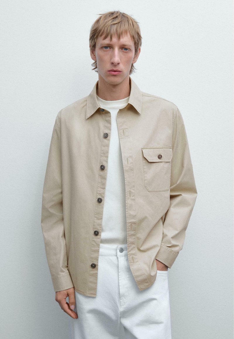 Beige skjorte med knapper lavet af bomuld, med en brystlomme, brune knapper og lange ærmer, parret med en hvid T-shirt med rund hals og hvide bukser.