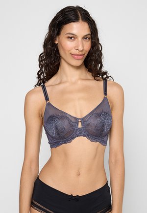 Kobieta o długich, kręconych włosach, ubrana w ciemny koronkowy biustonosz i czarne majtki z wysokim stanem ozdobione koronką, stojąca na jednolitym tle.