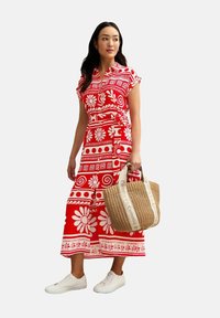 Vestido rojo con patrones florales blancos y diseños geométricos, mangas cortas, cintura atada, combinado con un bolso de paja beige y zapatillas blancas.
