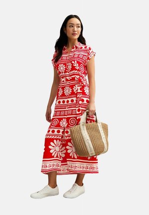Red dress with white floral patterns and geometric designs, short sleeves, tied waist, paired with a beige woven bag and white sneakers.
