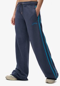 Persoon in donkerblauwe wijde sweatpants met turkooise zijstrepen, met een trekkoord taille en grijze sneakers, staand met de handen in de zakken.