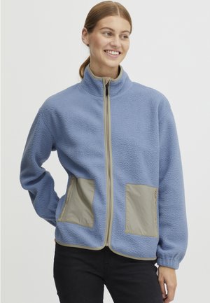 Blue fleece jacket with a high collar, a zip front, and two beige patch pockets. Features elastic cuffs and a textured fabric finish.