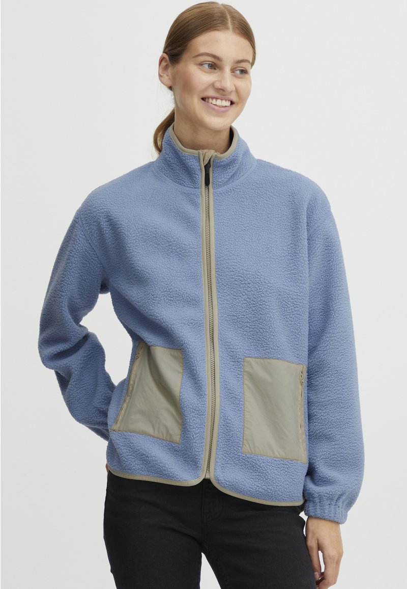 Blå fleecejacka med hög krage, dragkedja framtill och två beige fickor i tyg. Har elastiska ärmslut och en texturerad yta.