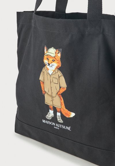 Μαύρη τσάντα tote με εικονογραφημένη αλεπού που φοράει μπεζ σύνολο και καπέλο, στέκεται με τα χέρια στις τσέπες. Κείμενο "Maison Kitsuné Paris" από κάτω.