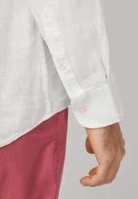 Witte linnen blouse met lange mouwen, voorzien van een geknoopte manchet en een relaxte pasvorm. De mouwen zijn iets opgerold, waardoor een gestructureerde stof zichtbaar is.