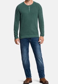 Zelená dlouhá rukávová henley košile s texturovanou tkaninou, kulatým výstřihem a třemi knoflíky, v kombinaci s modrými džíny a hnědými botami.