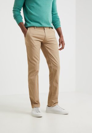 Uomo che indossa pantaloni beige, una maglia a maniche lunghe color teal, sneakers bianche, in piedi con le mani in tasca davanti a uno sfondo semplice.