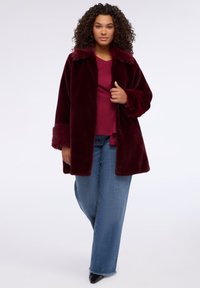 Burgundský plášť z umělé kožešiny s límcem a texturovanými rukávy, v kombinaci s třešňovým topem a širokými modrými džíny. Kabát má volný střih.