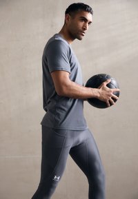 Szürke sport póló és szoros szürke leggings, egy fekete sportlabdát tartva, egy texturált világosszürke háttér előtt.