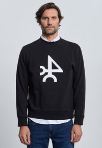 Hombre con sudadera negra con diseño geométrico abstracto blanco, camiseta blanca debajo y vaqueros azules, de pie frente a un fondo liso.