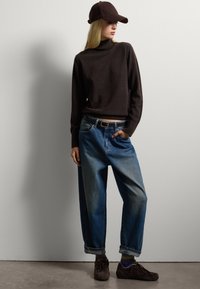 Maglione a collo alto marrone, jeans baggy azzurri chiari con orli arrotolati, sneaker scure e un berretto marrone. Design semplice e informale.