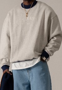 Šedá mikina s kulatým výstřihem a námořnicky modrými manžetami s vyšívaným textem "BLKVIS NY." Nositelná přes bílou košili a spárovaná se světle modrými džínami.