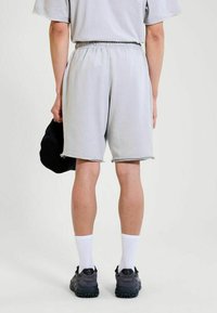 Persona vistiendo pantalones cortos elásticos de color gris claro, calcetines blancos hasta la altura del tobillo, zapatillas oscuras, sosteniendo una gorra negra, de pie frente a un fondo blanco.