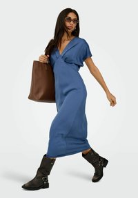 Niebieska sukienka kopertowa z krótkimi rękawami, gładka tkanina i zwiewny fason. W zestawie z czarnymi botkami za kostkę i brązową torbą na ramię.