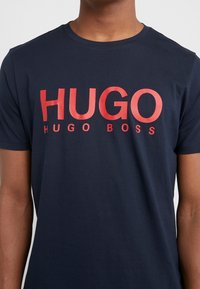 Tmavě modré bavlněné tričko s velkým červeným logo "HUGO" a menším textem "HUGO BOSS" pod ním, kulatým výstřihem a krátkými rukávy.