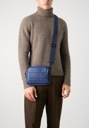 Borsa a tracolla rettangolare blu con dettagli trapuntati, realizzata in materiale liscio, indossata su un maglione marrone. Inclusa tracolla regolabile.